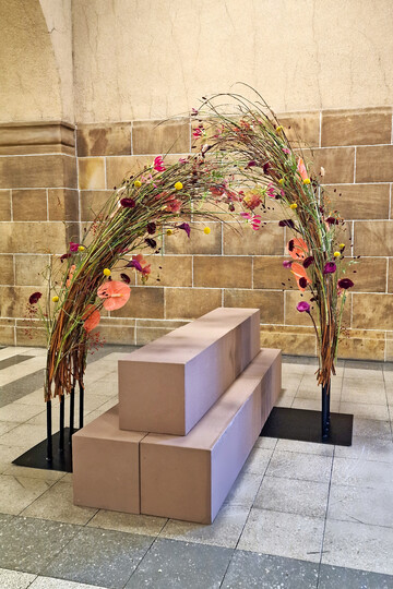 Die gebogene Anordnung, die den Sarg umgibt, stellt Bewegung und Freiheit dar � wie es auch bei einer T�nzerin auf der B�hne der Fall w�re. Zwei Metallplatten und darauf geschwei�te Rohre dienen als technische Basis. Die Weide dient sowohl als geschwungenes Gestaltungselement als auch als technische Basis f�r die Blumen. Eine freie Anordnung der passend ausgesuchten Bl�ten verst�rkt die Aussage des Sargschmucks.