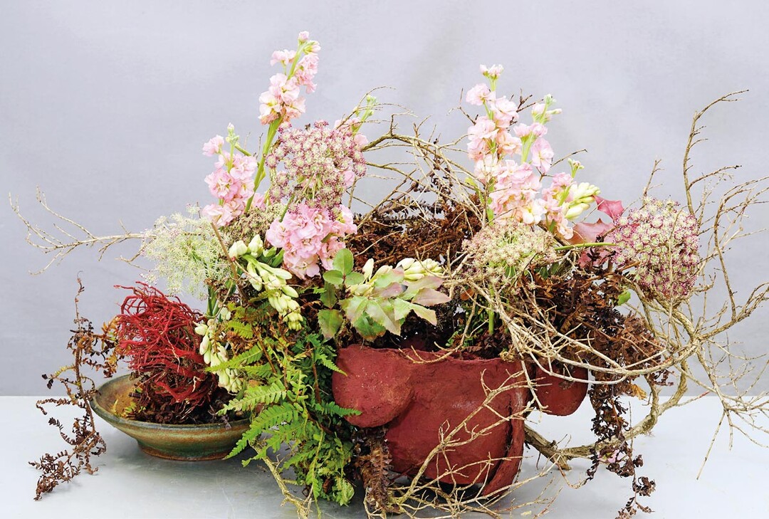 Eine Art spontanes Stillleben: Eine abgestorbene Pflanze mit ausgewaschenem Wurzelballen ist fest mit einer Form verbunden, die kein Behältnis darstellt, sondern ohne Boden offen daliegt. Sie ist aus einer Ton- Papier-Masse hergestellt und wirkt ein wenig wie unter einem Strauch vergessen. Passende Kontraste liefern Farn, Levkojen, Ammi …